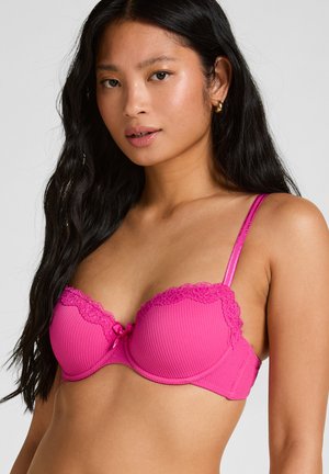 Jeune femme aux longs cheveux noirs portant un soutien-gorge rose vif orné de dentelle sur un fond gris uni.