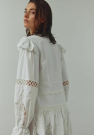 Balta kleita ar izšūtām mežģīnēm uz piedurknēm, acs raksta volāniem uz pleciem un daudzpakāpju svārkiem ar dekoratīvām šuvēm.