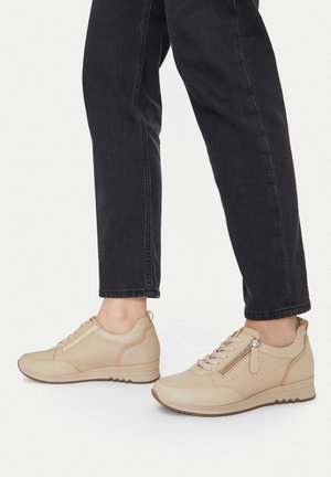 Beige ādas sporta apavi ar perforētu dizainu, priekšējo šņorēšanu, sānu rāvējslēdzēju un teksturētu gumijas zoli. Valkāti ar tumšām džinsām.