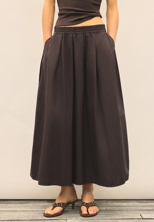Personne portant une jupe noire taille haute, large avec poches et des sandales à talons noires, debout sur un sol en bois.