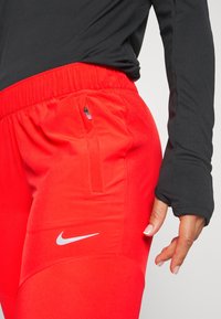 Rode sportbroek met een elastische tailleband en een zijzak met rits, gecombineerd met een zwart shirt met lange mouwen en aansluitende mouwen. Zilveren Nike-logo.