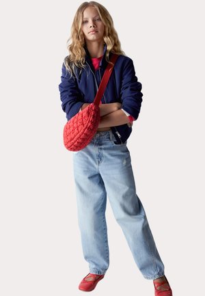 Borsa a tracolla imbottita rossa, giacca bomber blu, camicia rossa, jeans baggy azzurri chiari e scarpe flat rosse con cinture incrociate.