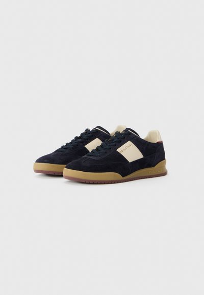 Et par marineblå mokkasinsneakers med beige sidepaneler, gummisåler og sorte snørebånd på hvit bakgrunn.