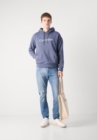 Felpa grigia con logo "Calvin Klein", tasca frontale, abbinata a jeans azzurri e sneakers bianche, mentre tiene una borsa tote beige.