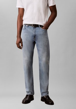 Mand iført lyseblå jeans med lige ben, sort bælte, hvid t-shirt og sorte lædersko, stående foran en ensfarvet grå baggrund.