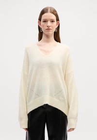 Pullover oversized color crema chiaro con scollo a V, spalle cadenti, lavorazione a maglia testurizzata e orlo che scende sul retro. Indossato con pantaloni neri.
