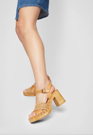 Jambes portant des sandales à plateforme beige avec des brides tressées, des clous et une boucle, accompagnées d'une jupe en denim courte sur fond uni.