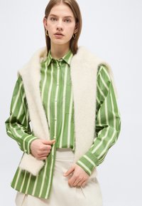 Νεαρή γυναίκα που φορά χρυσούς κρίκους στα αυτιά, πουκάμισο με πράσινες και λευκές ρίγες, κρεμ φούστα και άσπρο χνουδωτό γιλέκο ριγμένο στους ώμους.