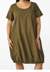 Mujer con vestido suelto de manga corta color verde oliva con dobladillo fruncido y bolsillos laterales, de pie con las manos relajadas a los lados.