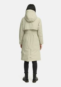 Lange beige gewatteerde jas met een capuchon, voorzien van een elastische tailleband en een korter achterkantontwerp. Draagt over donkere spijkerbroeken en zwarte laarzen.
