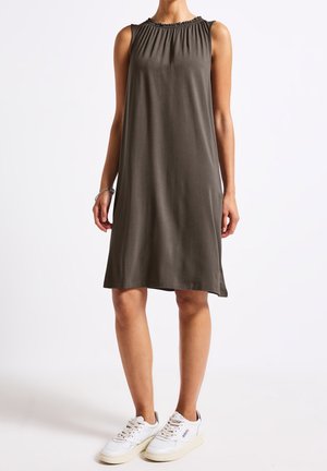 Kobieta ubrana w bezrękawnikową, ciemnooliwkową sukienkę do kolan i białe trampki, stoi na tle jednolitego jasnego tła.