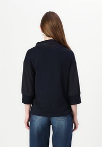 Granatowy sweter z wysokim kołnierzem i rękawami trzyczwartowymi, o gładkiej fakturze i luźnym kroju, w połączeniu z jasnoniebieskimi dżinsami.