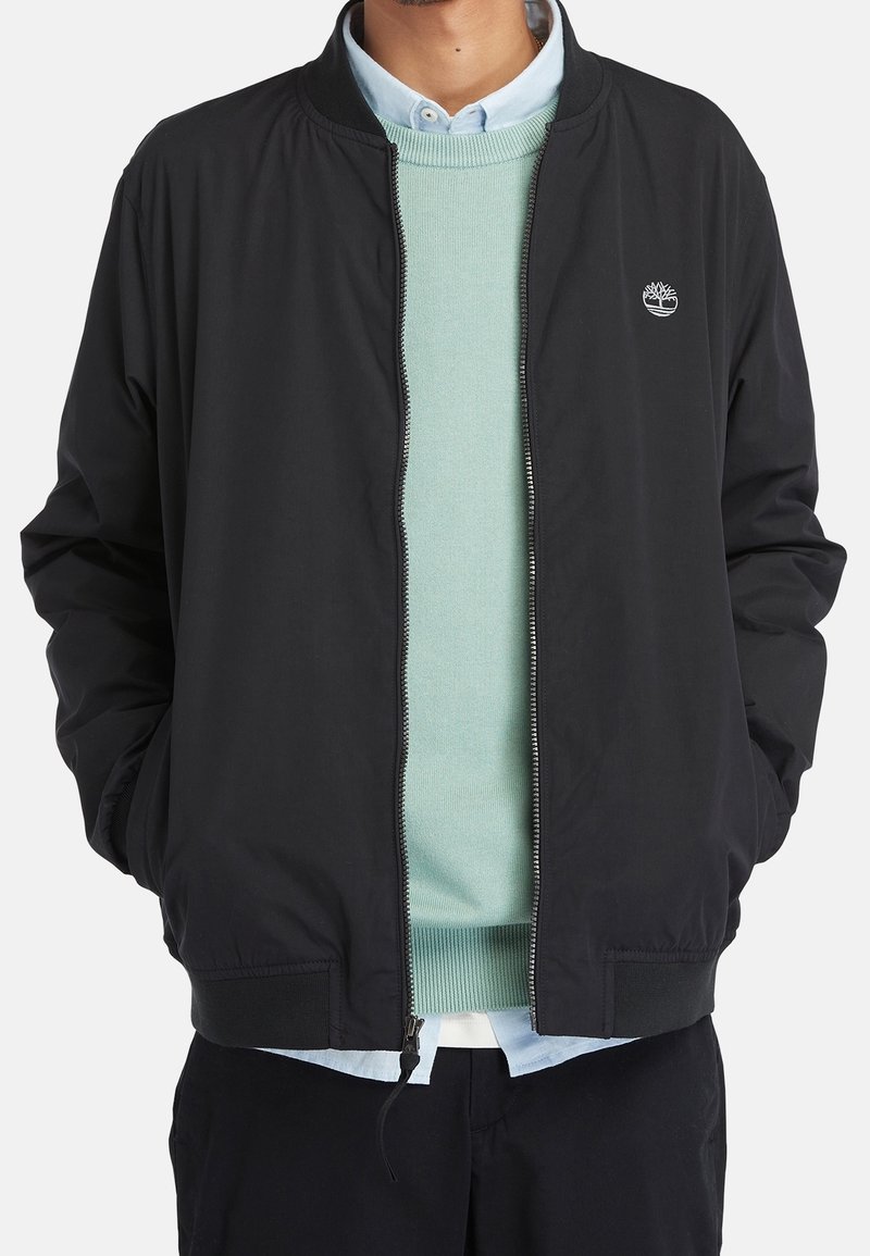 Homme portant une veste noire zippée sur un pull vert menthe et une chemise bleu clair à col, mains dans les poches, fond blanc.
