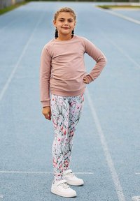 Jasnoróżowa koszula z długim rękawem i białe legginsy z kwiatowym wzorem, połączone z białymi trampkami. Stoi na niebieskiej bieżni lekkoatletycznej.