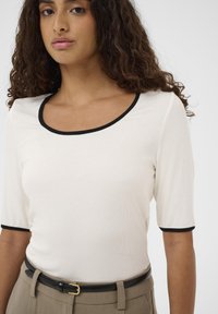 Tricou crem cu dungi, cu mâneci scurte, margine neagră în jurul gâtului rotund și al mânecilor, purtat cu o curea neagră și pantaloni bej.