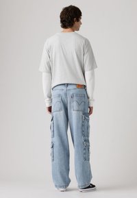 Ανοιχτό γκρι κοντομάνικο μπλουζάκι με στρογγυλή λαιμόκοψη, φορεμένο πάνω από λευκό μακρυμάνικο πουκάμισο, συνδυασμένο με φαρδιά μπλε τζιν παντελόνια cargo με πολλές τσέπες.