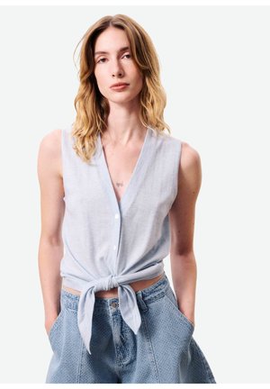 Jeune femme aux cheveux blonds ondulés portant un haut sans manches bleu clair boutonné, noué à la taille, et un pantalon en jean texturé taille haute.