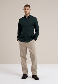Donkergroene button-up shirt met een slim fit, gecombineerd met lichtbeige broek. Het shirt heeft een klassieke kraag en lange mouwen, gedragen met zwarte schoenen.