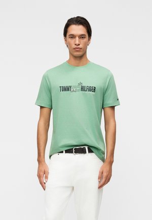 Junger Mann trägt ein grünes Tommy Hilfiger T-Shirt mit weißen Hosen und schwarzem Gürtel, steht vor einem einfarbigen hellgrauen Hintergrund.
