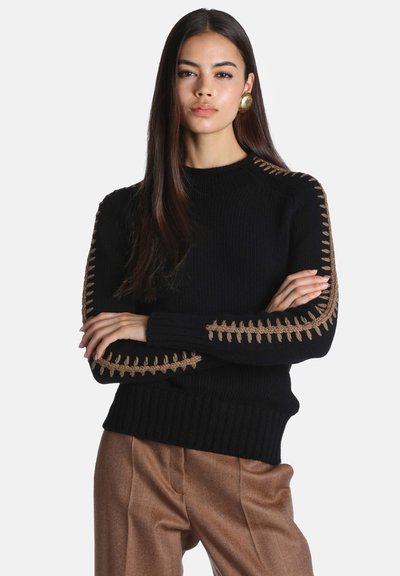 Jeune femme aux longs cheveux foncés, portant un pull noir avec des détails marron cousus sur les manches et un pantalon marron, bras croisés.
