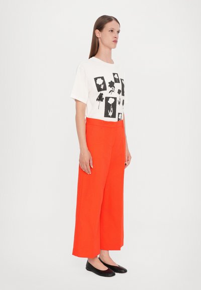 Hvid t-shirt med sorte blomsterprints, parret med højttaljede, vide orange bukser. Sorte flade sko fuldender outfittet.