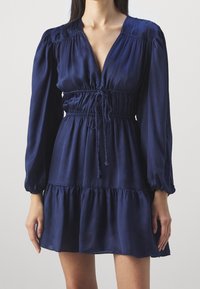 Námorně modrá saténová šaty s hlubokým V-výstřihem, nabíranými rukávy, nařaseným páskem a volánkovým lemem s nastavitelnými detaily na stahování.