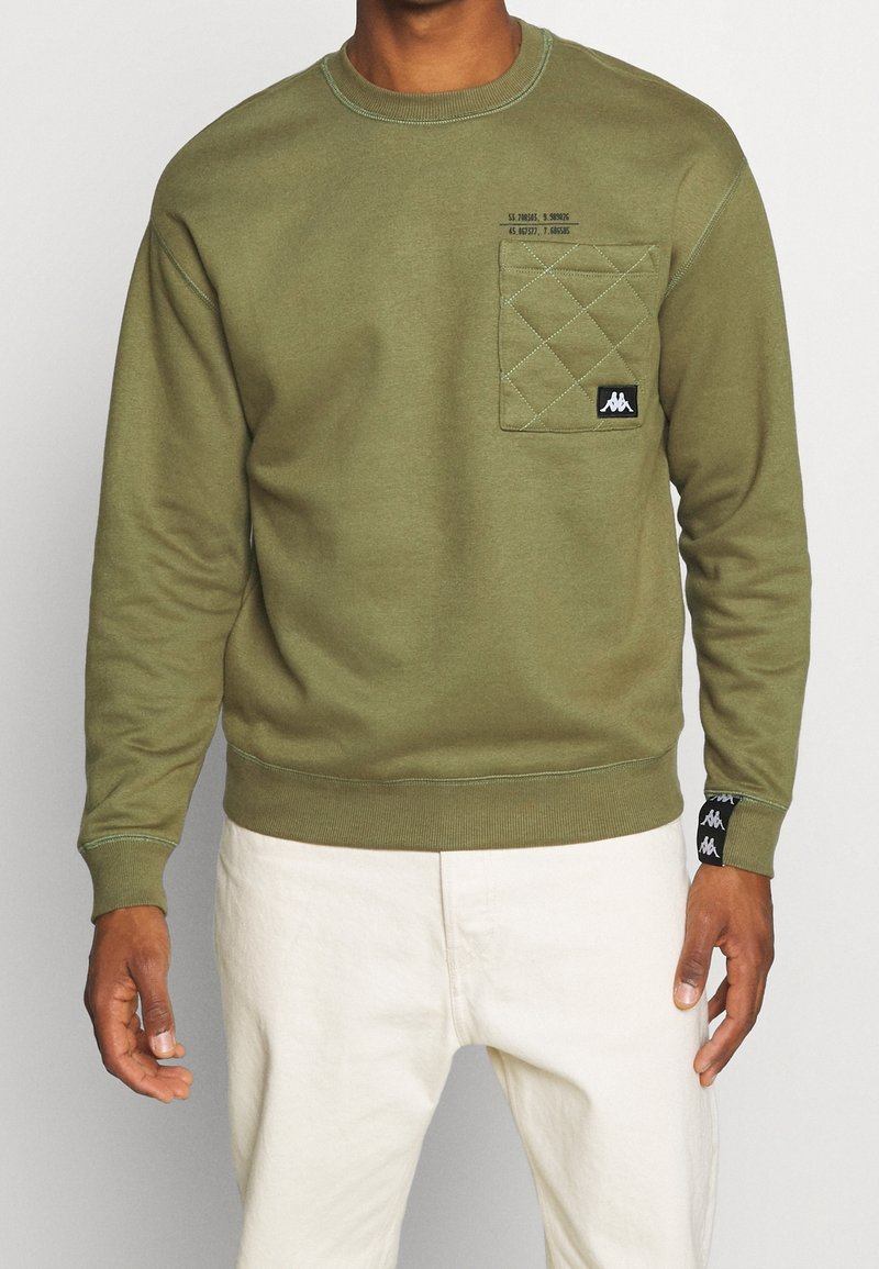 Homme portant un sweat matelassé vert olive avec poche avant et écussons de logo, associé à un pantalon blanc cassé, debout devant un fond uni.