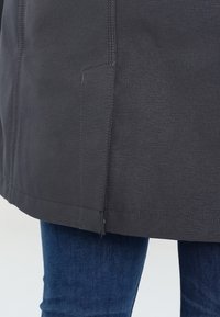 Tummanharmaa takki, jossa on teksturoitu pinta, suora helma ja sivusaumataskut, yhdistettynä sinisiin farkkuihin.