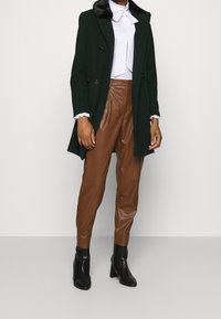 Cappotto doppiopetto verde scuro con colletto in pelliccia sintetica, indossato su una camicetta bianca con polsini arricciati, abbinato a pantaloni in pelle sintetica marrone e stivaletti neri.