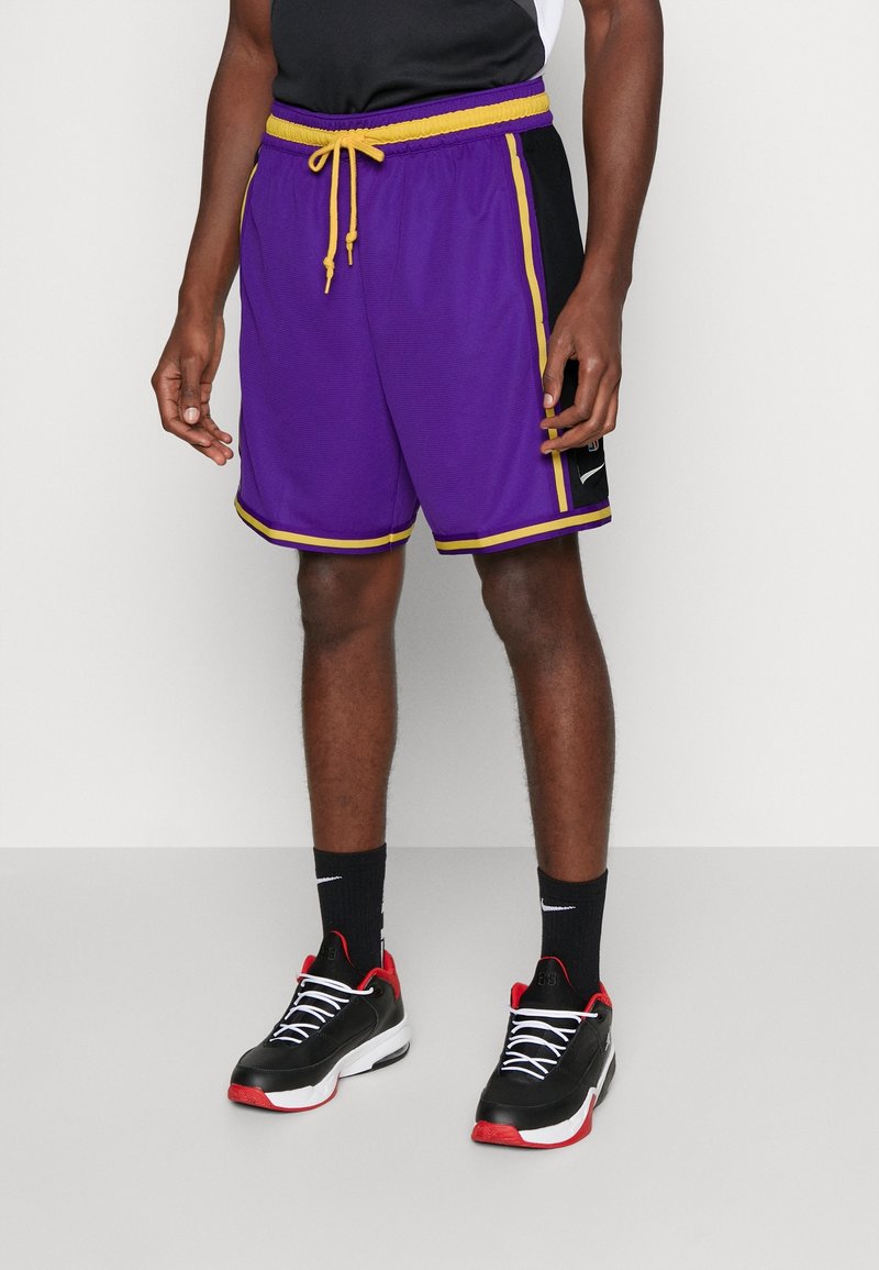 Shorts de basket violets avec des côtés noirs, cordon jaune et bordure, dotés d'un tissu texturé et de poches latérales. Associés à des baskets noires et rouges.