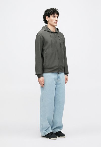 Γκρι φούτερ με φερμουάρ και υφασματικό μοτίβο, σε συνδυασμό με ανοιχτομπλε τζιν παντελόνι φαρδιάς γραμμής και μαύρα αθλητικά παπούτσια, φορεμένα από έναν άνδρα που στέκεται μπροστά σε λευκό φόντο.