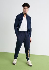 Námornícky zipsový sveter cez biely tričko, v kombinácii s námorníckymi teplákmi so značkou "Timberland" a svetlými teniskami.