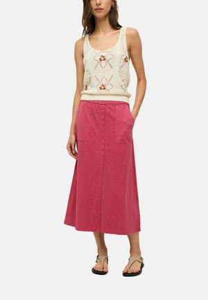 Mujer que lleva un top sin mangas de punto color crema con diseño floral, una falda midi rosa con botones delanteros y bolsillos, y sandalias beige.