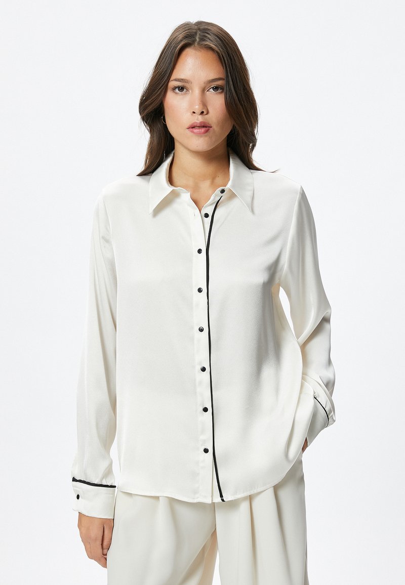 Camisa de seda blanca con ribetes negros, frontal con botones, mangas largas y botones negros. Textura suave y diseño de cuello clásico.