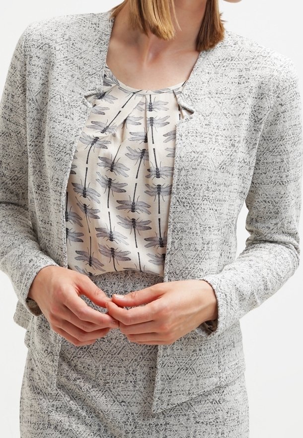 Femme portant un tailleur blazer texturé gris clair et une jupe, avec un chemisier beige imprimé de libellules noires, mains jointes à la taille.