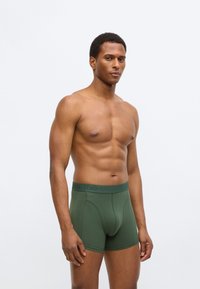 Πράσινα βαμβακερά μποξεράκια με ελαστική μέση που φέρει ανάγλυφα γράμματα. Η εφαρμογή είναι στενή και το σχέδιο περιλαμβάνει υποστήριξη με θήκη.