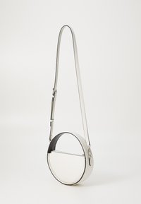 Rund crossbody-bag i hvitt med en svart aksent, laget av glatt lær. Har en glidelåslomme og en justerbar stropp.