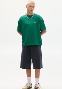 Tricou scurt verde, cu logo discret, asociat cu pantaloni scurți gri închis și adidași bej cu accente maronii. Fundal alb simplu.