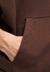 Ύφασμα καφέ φούτερ που δείχνει υφή, με μια τσέπη στο μπροστινό μέρος. Ένα χέρι έχει εισαχθεί στην τσέπη, αναδεικνύοντας την απαλότητα του υλικού.