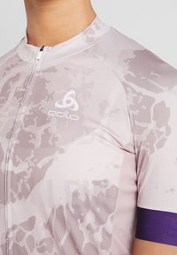 Ljusrosa cykeltröja med camomönster, korta ärmar, en fjärdedels dragkedja och lila detaljer på ärmen. Varumärkeslogotyp synlig.