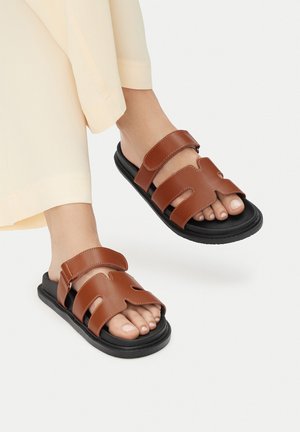 Brązowe skórzane sandały z dwoma szerokimi paskami, ozdobione wycięciami. Czarne gumowe podeszwy zapewniają amortyzację. W tle jasne spodnie.
