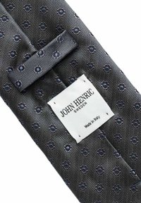 Harmaa kuvioitu solmio, jossa on pieniä sinisiä kukkakuvioita ja valkoinen etiketti, jossa lukee John Henric Sweden, valmistettu Italiassa.