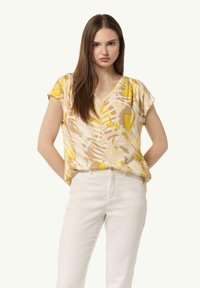 Kurze, V-Ausschnitt Bluse in cremigem Beige mit abstrakten gelben und braunen Mustern. Kombiniert mit hellen, figurbetonten Hosen.