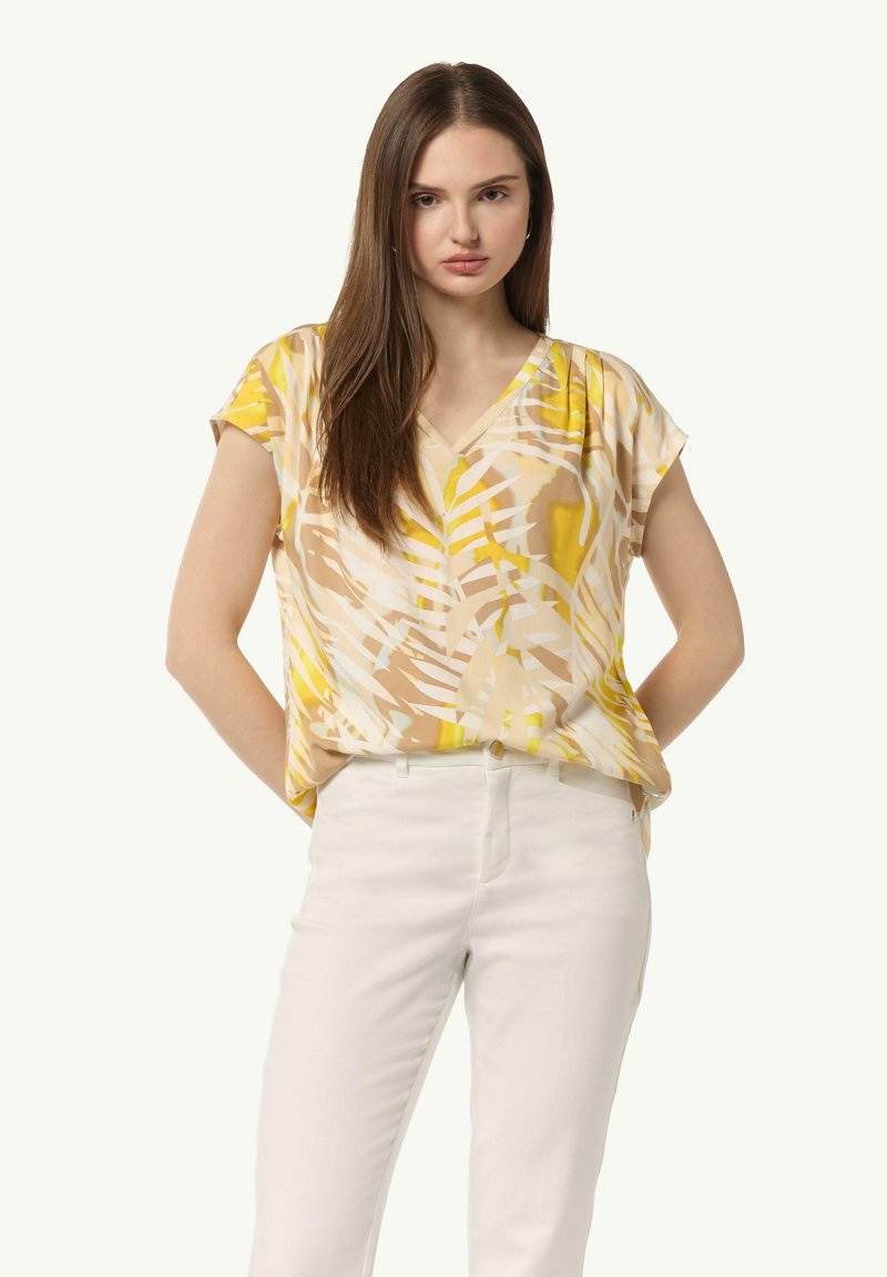 Kurze, V-Ausschnitt Bluse in cremigem Beige mit abstrakten gelben und braunen Mustern. Kombiniert mit hellen, figurbetonten Hosen.