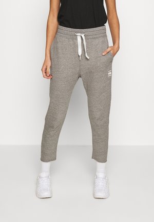 Person iført grå joggingbukser med snore, hvide sneakers og hvide sokker, med hænderne afslappet ned langs siderne mod en ensfarvet baggrund.