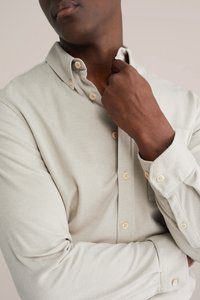 Šviesiai pilka dryžuota marškinėliai su sagomis ant apykaklės, šviesios spalvos sagutės ir minkšta tekstūra; ilgi rankovės suktos prie rankogalų.