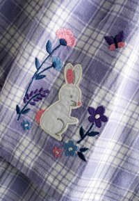 Patch en forme de lapin blanc entouré de fleurs brodées roses, violettes et bleues, ainsi que d'un papillon, sur un tissu à carreaux violet et blanc.