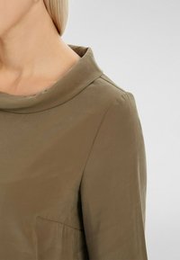 Blouse en satin vert olive avec un col drapé, des manches longues et une texture subtile. Dotée d'une petite poche sur le côté gauche.