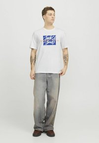 Witte t-shirt met korte mouwen met een blauw en oranje "Jack & Jones Essentials" graphic. Gepaard met lichtgewassen brede jeans en bruine schoenen.