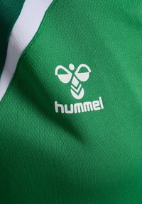 Zielona sportowa tkanina z białym logo Hummel i symbolem pszczoły w kształcie chevronu, ukazująca teksturę i szew z białym detalem.